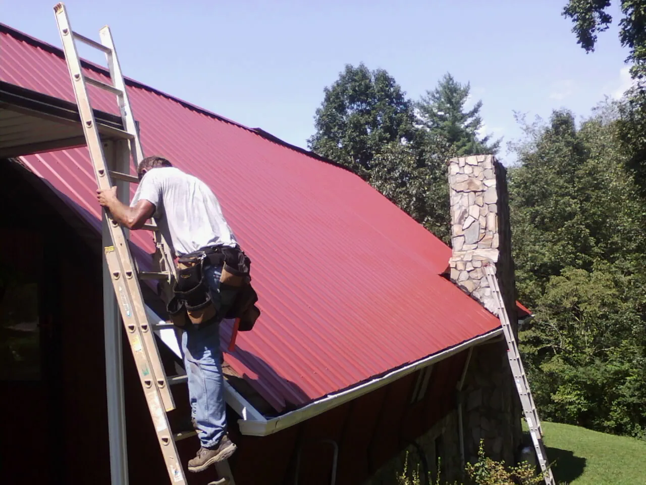 Roofer on a ladder beside a red metal roof and stone chimney — Soffit & Fascia Repair by Roofing Brothers in East Brunswick